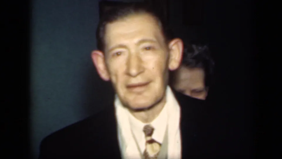 A 1949 New York home-movie frame shows a man in a dark suit and tie looking toward the camera inside a teal-walled interior.