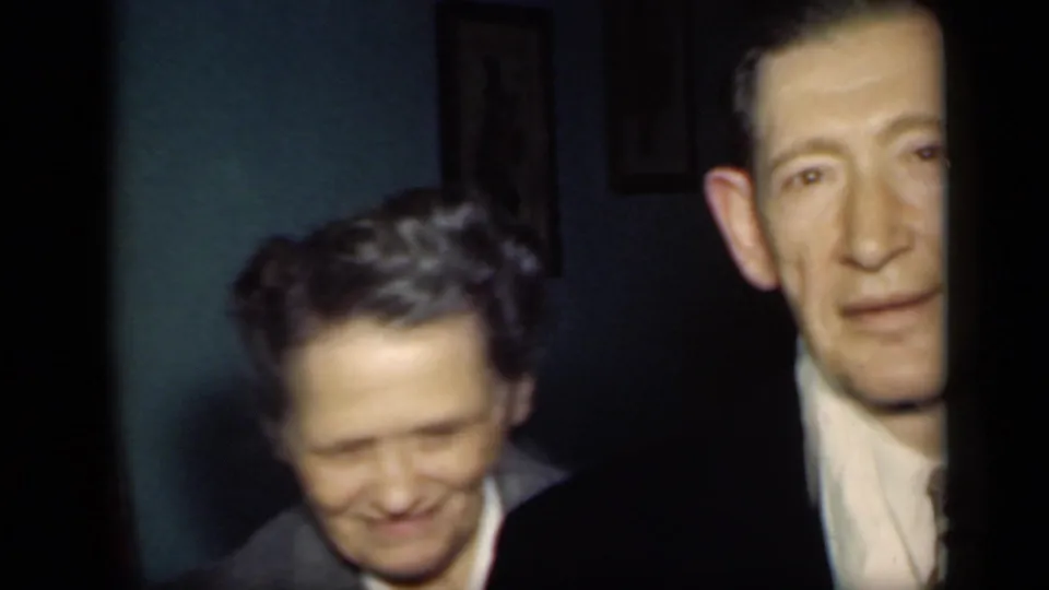 Two men appear in a close indoor 8mm frame from 1949 New York, with one smiling and the suited man partly cut off at the right edge.