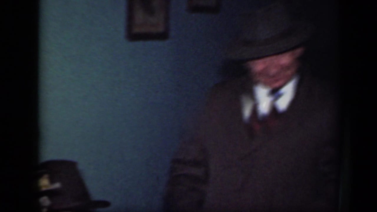 Man in a dark suit and fedora standing in a dim interior near a teal wall.