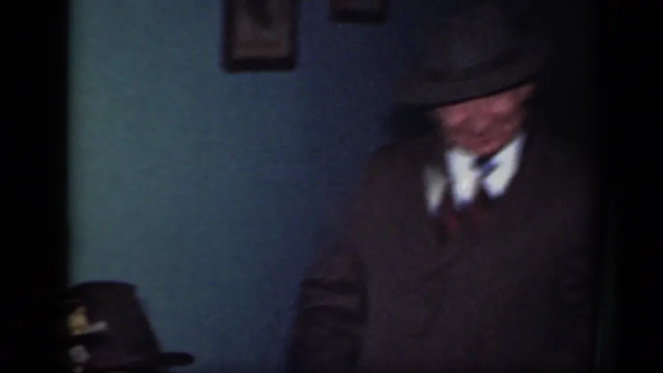 In a 1949 New York 8mm frame, a suited man in a fedora stands inside a dim room with framed wall art nearby.