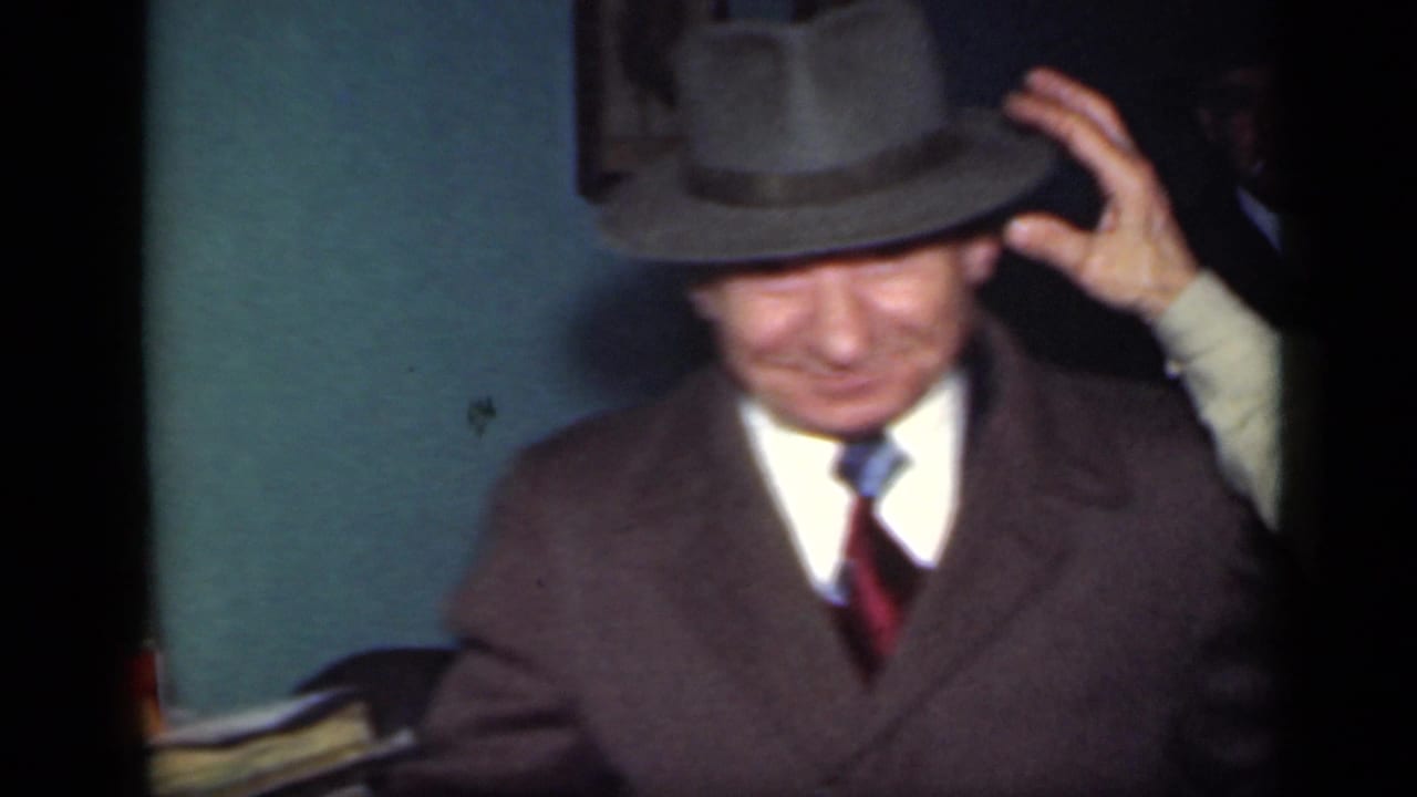A suited adult man smiles while lifting his fedora in a dim 1949 New York interior with a teal wall behind him.