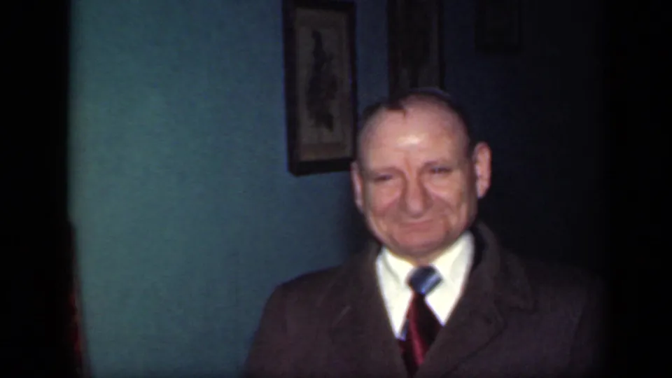 A balding man in a suit and tie smiles toward the camera in a 1949 New York home-movie interior.