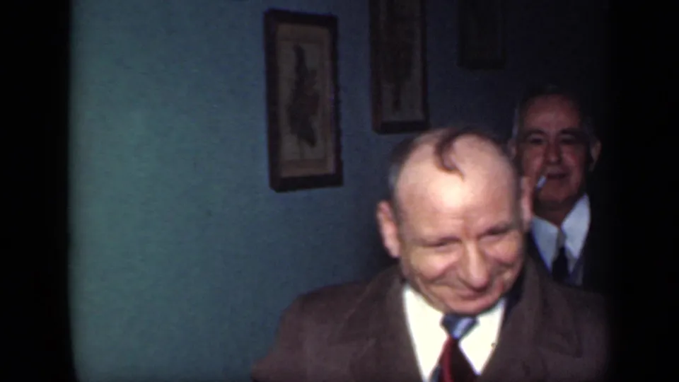 A smiling suited man stands in the foreground while another adult man appears behind him in the dim 1949 New York interior.