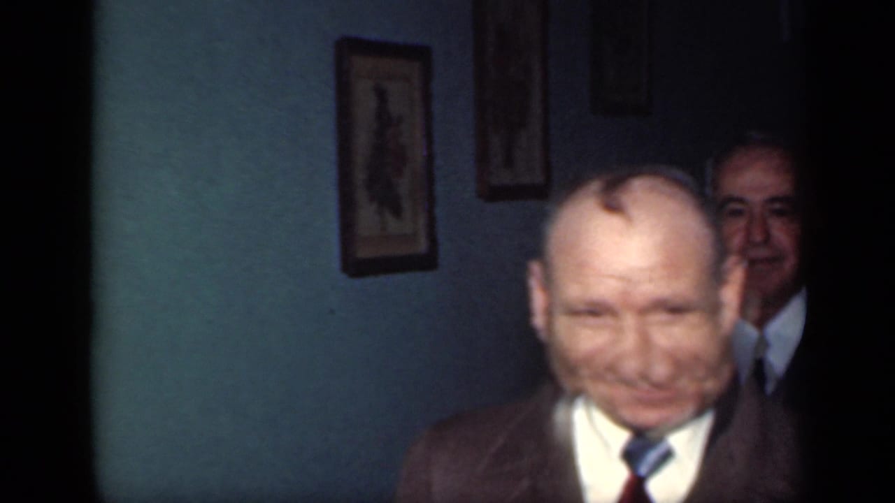 Two suited older men in a dim room, one close to camera and another partly behind him.