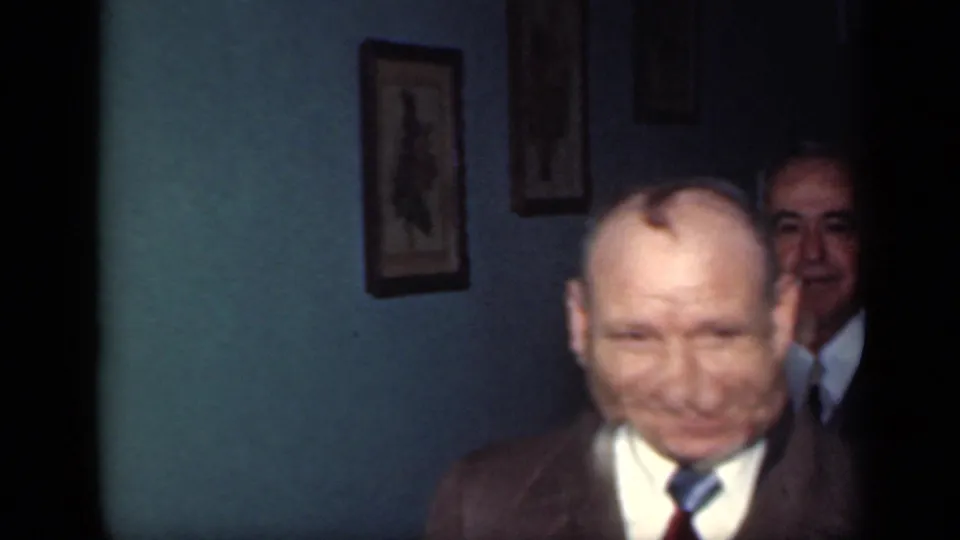 1949 New York home-movie frame of two older men in suits inside a dim teal-walled room with framed pictures.