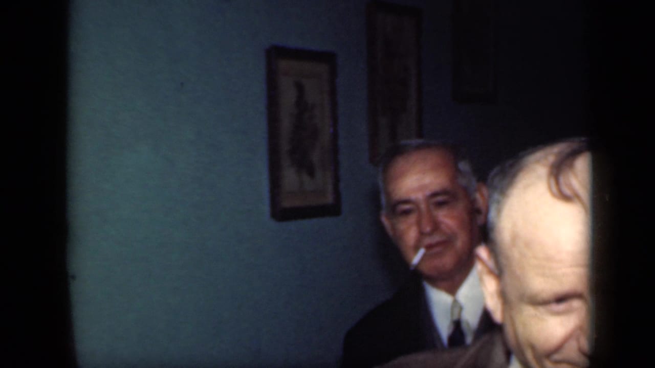 Older man in a suit holds a cigarette while another man's head crosses the foreground.