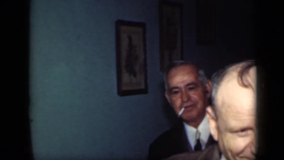 1949 New York interior frame with a suited older man holding a cigarette against teal walls and framed artwork.