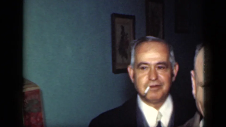 1949 New York home-movie frame showing an older suited man with a cigarette in a dim private-looking room.