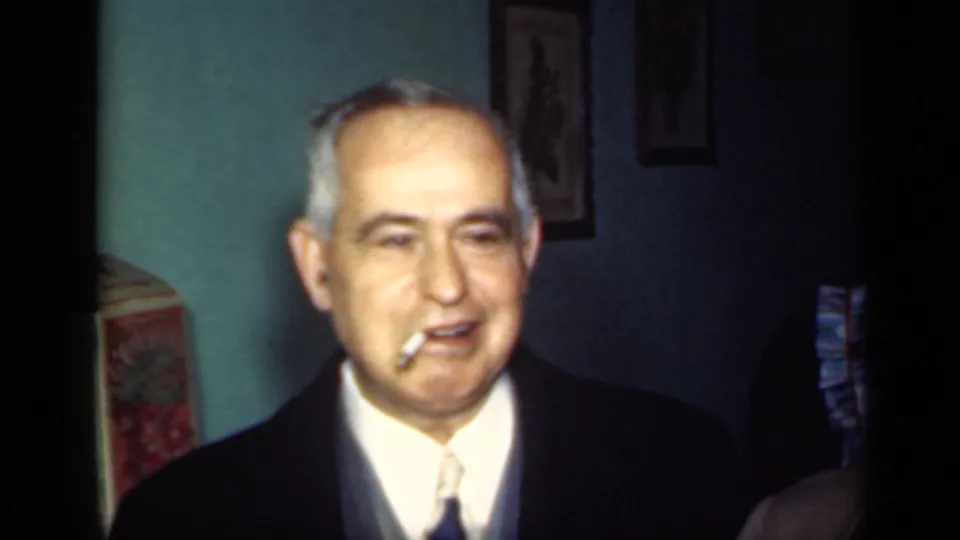1949 New York frame of a suited older man with a cigarette, lit by home-movie exposure in a teal-walled room.