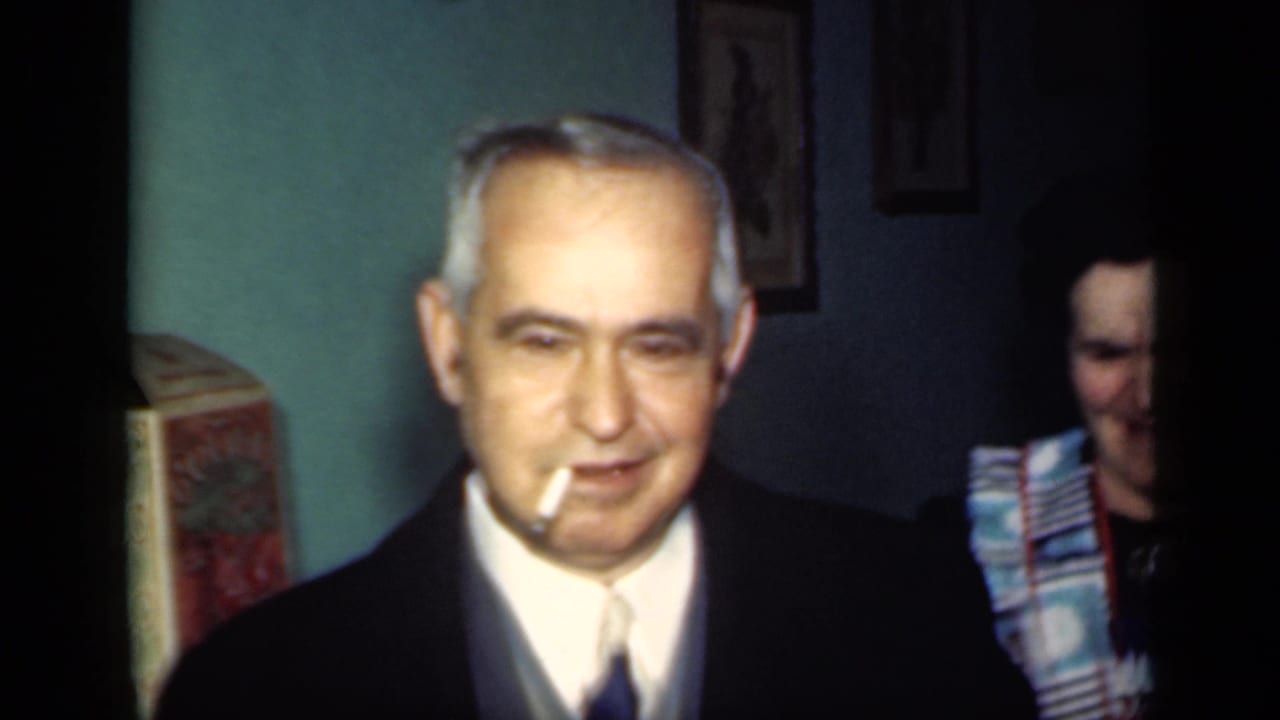 Older man in a dark suit smiles with a cigarette in his mouth inside a dim room.