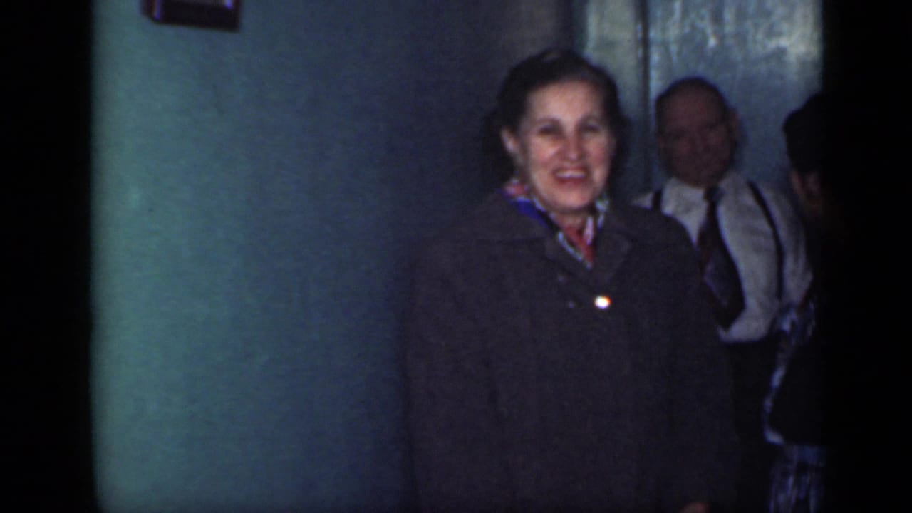 Older woman in a dark coat smiles near a doorway with other adults behind her.