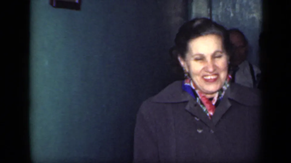 The same older woman smiles broadly in a dark coat and colorful scarf inside the dim 1949 New York home-movie interior.