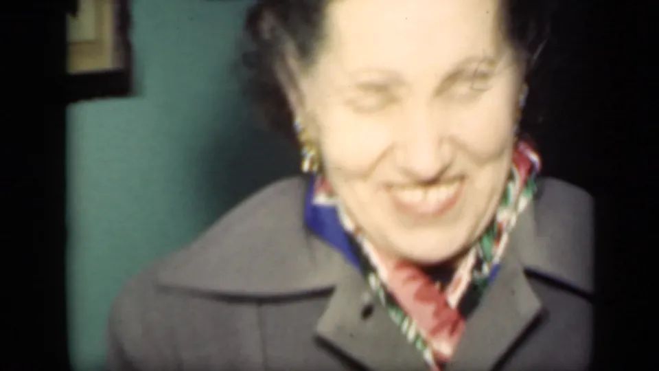 Close handheld frame of an older woman smiling in a coat and colorful scarf, with soft exposure from the 8mm New York home movie.
