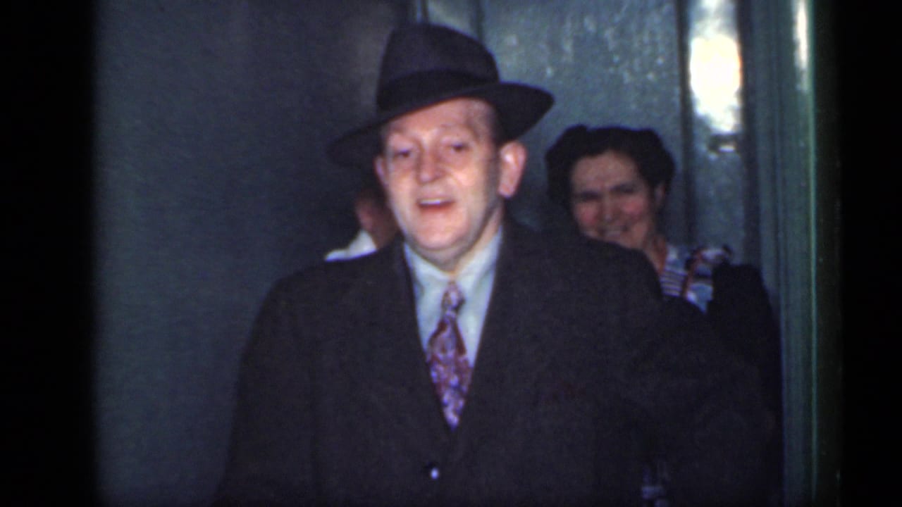 Man in a fedora and dark coat steps through a doorway with people behind him.