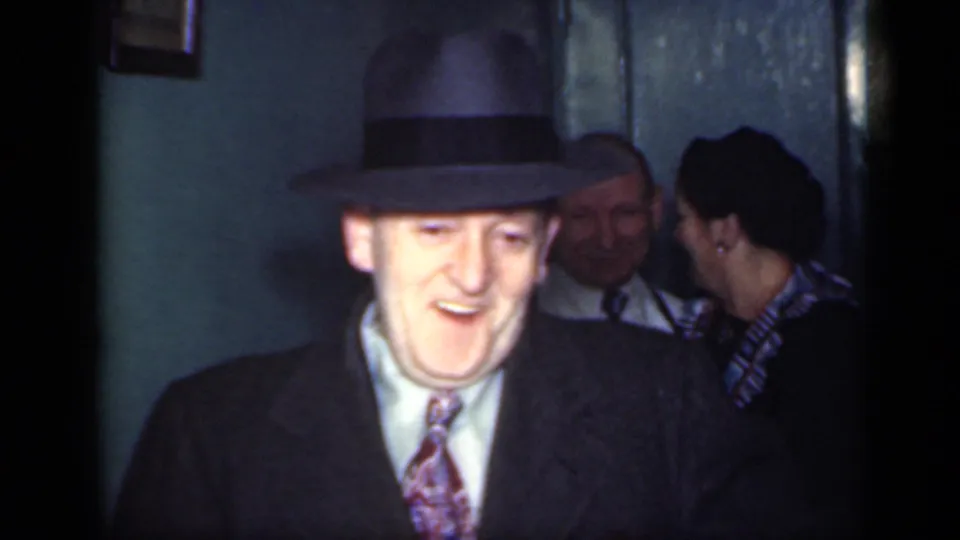 A smiling man in a fedora, dark coat, shirt, and tie faces the camera in this 1949 New York interior home-movie frame.