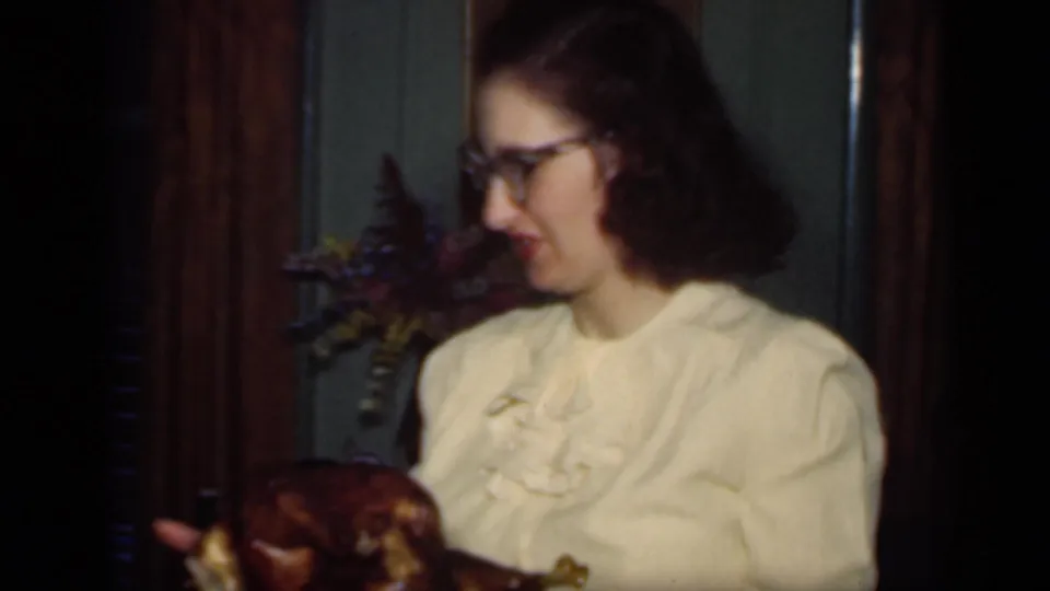 Young woman in glasses and a light blouse holds a roasted turkey in a private interior, matching the supplied 1949 New York metadata.
