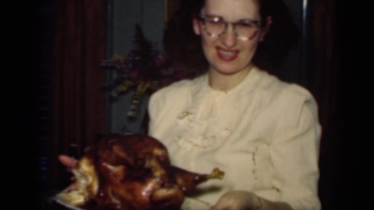 In 1949 New York home-movie footage, a woman in glasses and a cream blouse presents a roasted turkey on a platter indoors.