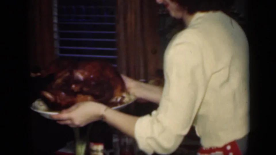 The woman, seen mostly in profile, steadies the roasted turkey platter in a dim private interior with window blinds behind her.