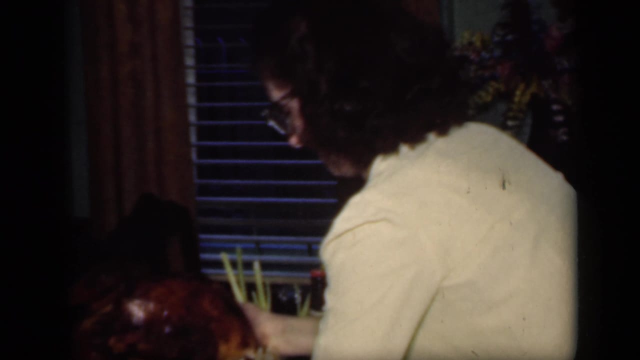 Woman in glasses leans over a dining table near a roasted turkey and tall greenery.