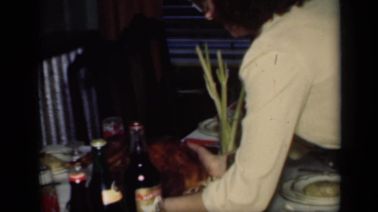 A woman in a cream sweater sets a roasted turkey on a formal dinner table with beer bottles, dishes, and patterned glassware visible.