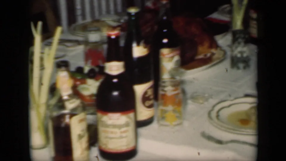 The frame centers on the set dining table, showing a roast turkey, several beer bottles, patterned tumblers, plates, and tall greenery in a vase.