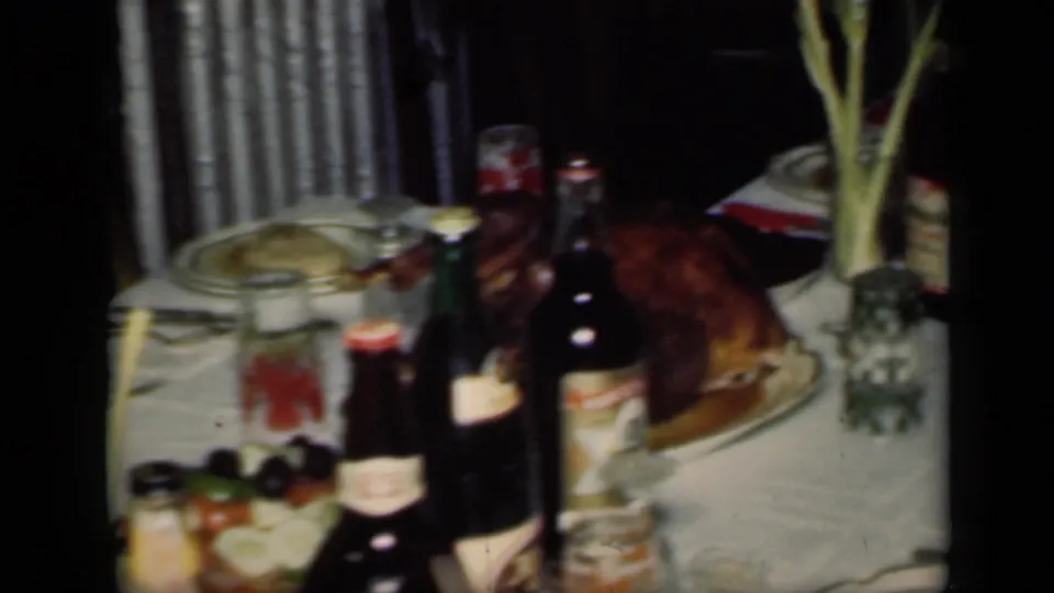 1949 New York home-movie frame showing the dinner table after serving, with bottles, jars, glasses, a roasted dish, and tall stems in a vase.