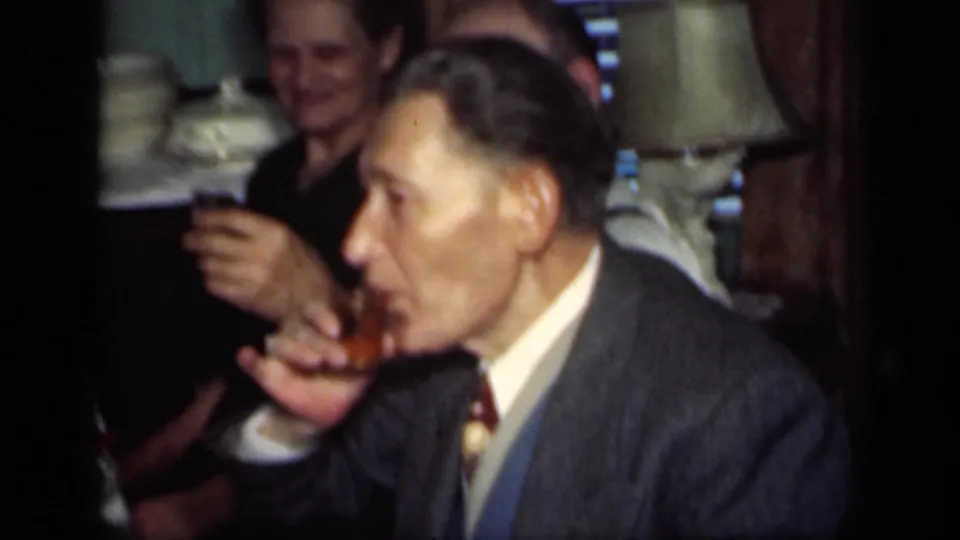 1949 New York home-movie frame of an older man in a suit sipping from a small glass, with another adult smiling in the background.