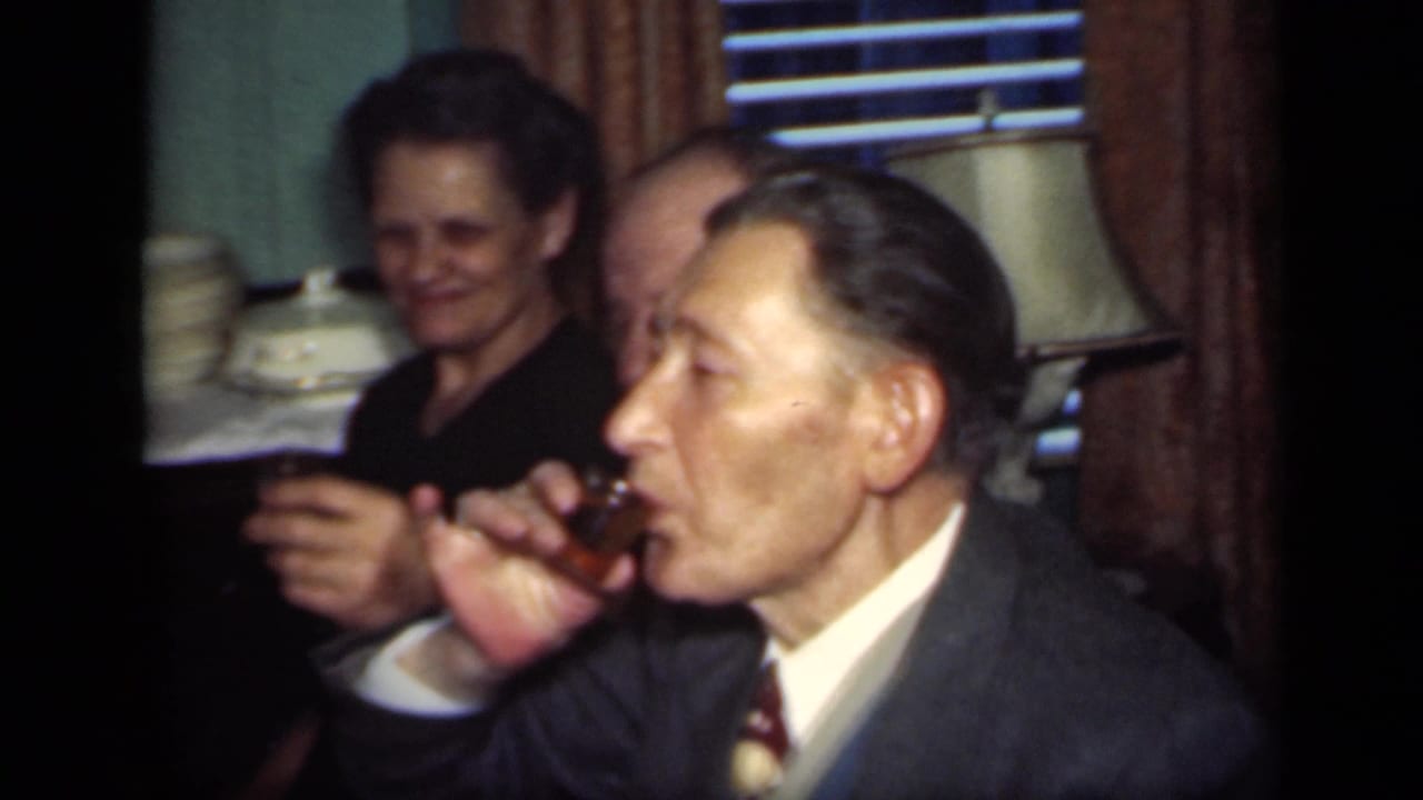 Man in a suit drinks from a small glass while seated near other adults at a table.