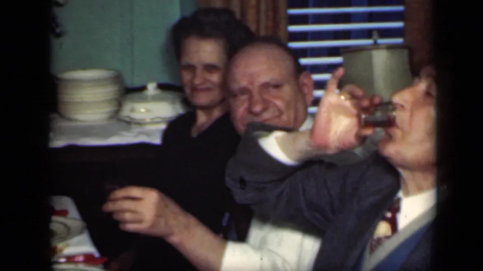 Adults at a New York dinner party hold small glasses, with one man lifting his drink and a woman smiling behind him.