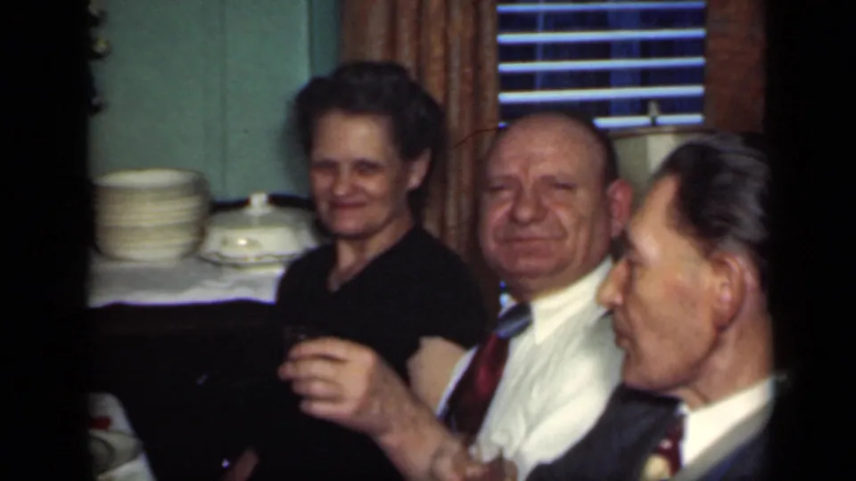 A woman smiles toward the camera while two men sit nearby during a 1949 New York indoor dinner gathering.