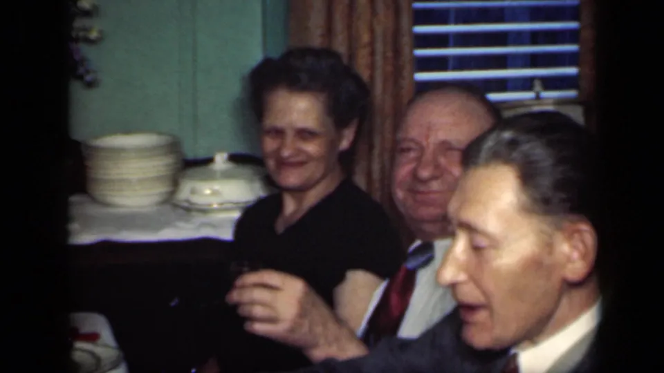 A smiling woman and seated man remain at the table as a suited man passes close to the camera in this home-movie frame.