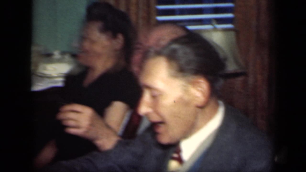Adults seated at a dining table, with a man in a suit holding a drink near a woman in a dark dress.