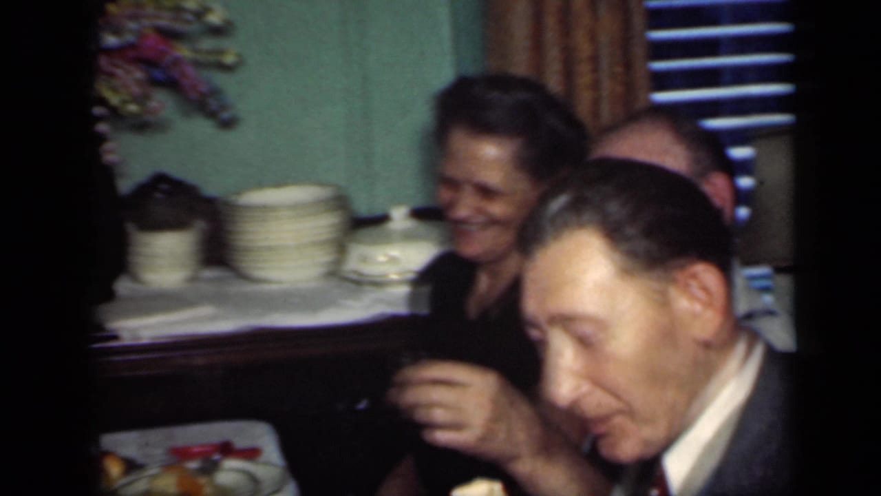 1949 New York dinner-party frame with a smiling woman, a suited man holding a drink, stacked plates, curtains, and blinds behind them.
