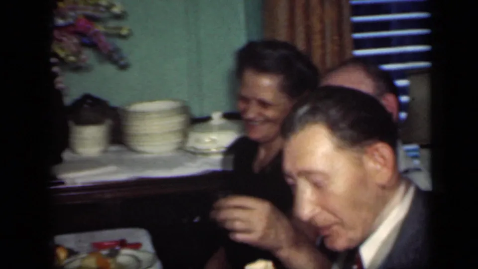 1949 New York dinner-party frame with a smiling woman, a suited man holding a drink, stacked plates, curtains, and blinds behind them.