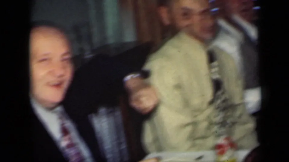 1949 New York home-movie frame showing men in suits around a dining table, with motion blur and party glassware visible.