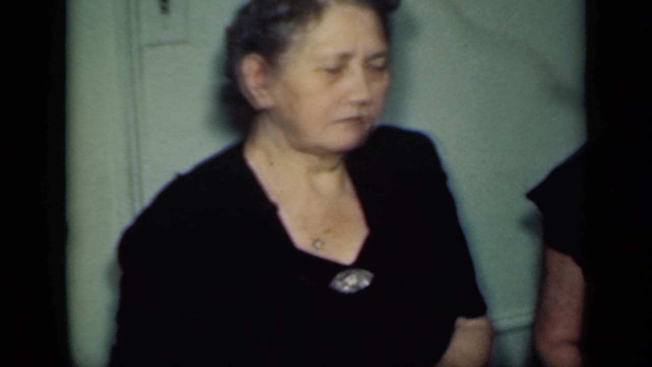 An adult woman in a black dress and brooch stands indoors against a green wall in the 1949 New York home-movie scene.