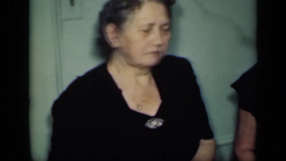 An adult woman in a black dress and brooch stands indoors against a green wall in the 1949 New York home-movie scene.