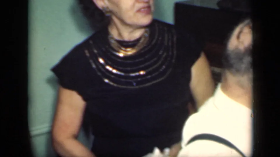 A woman wearing a black dress and gold necklace stands indoors near a bearded man in profile in the 1949 New York clip.