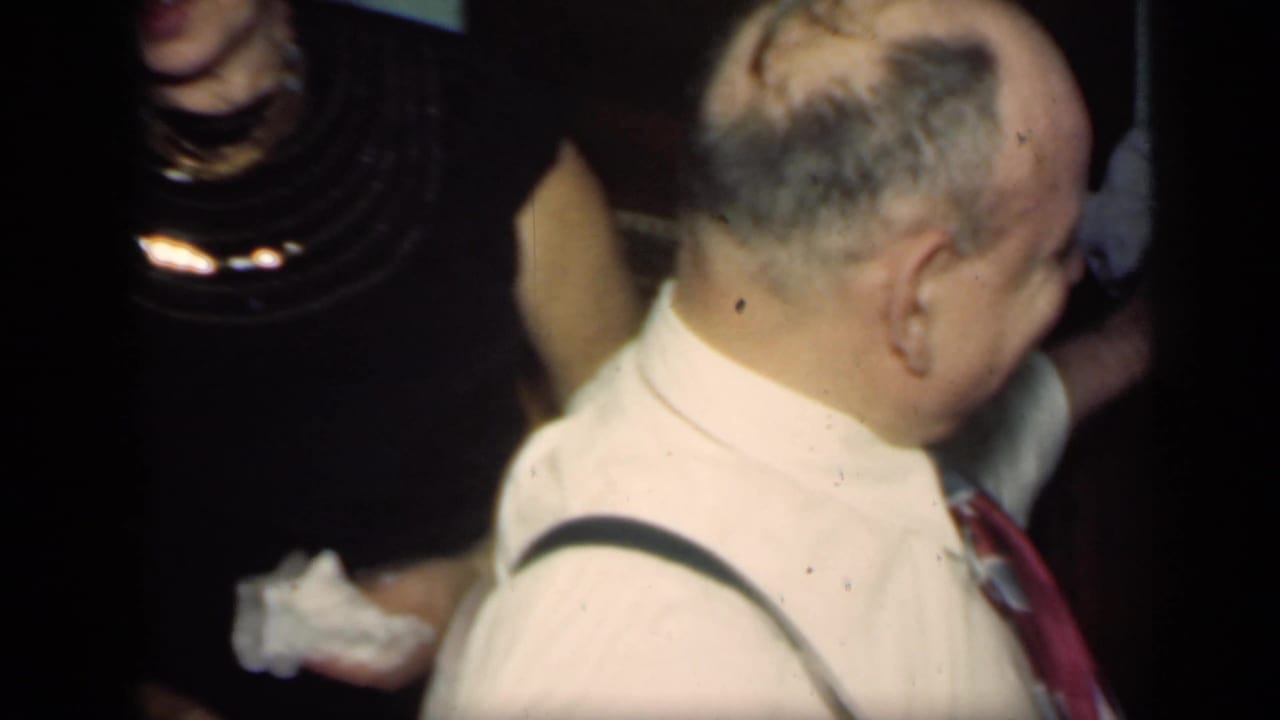 Adult man in suspenders turns toward others at a dim indoor party.