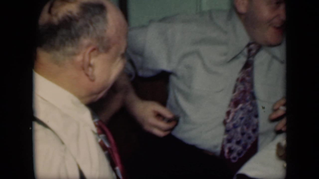 A man in a light shirt and suspenders turns toward a smiling man in a patterned tie inside a crowded New York room.