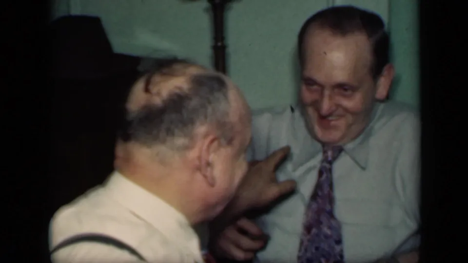 In a private indoor gathering, two adult men smile and gesture toward each other, with one man wearing suspenders and the other a patterned tie.