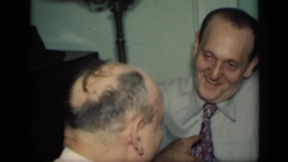 A smiling man in a patterned tie looks toward another man seen from behind during a 1949 New York indoor social scene.