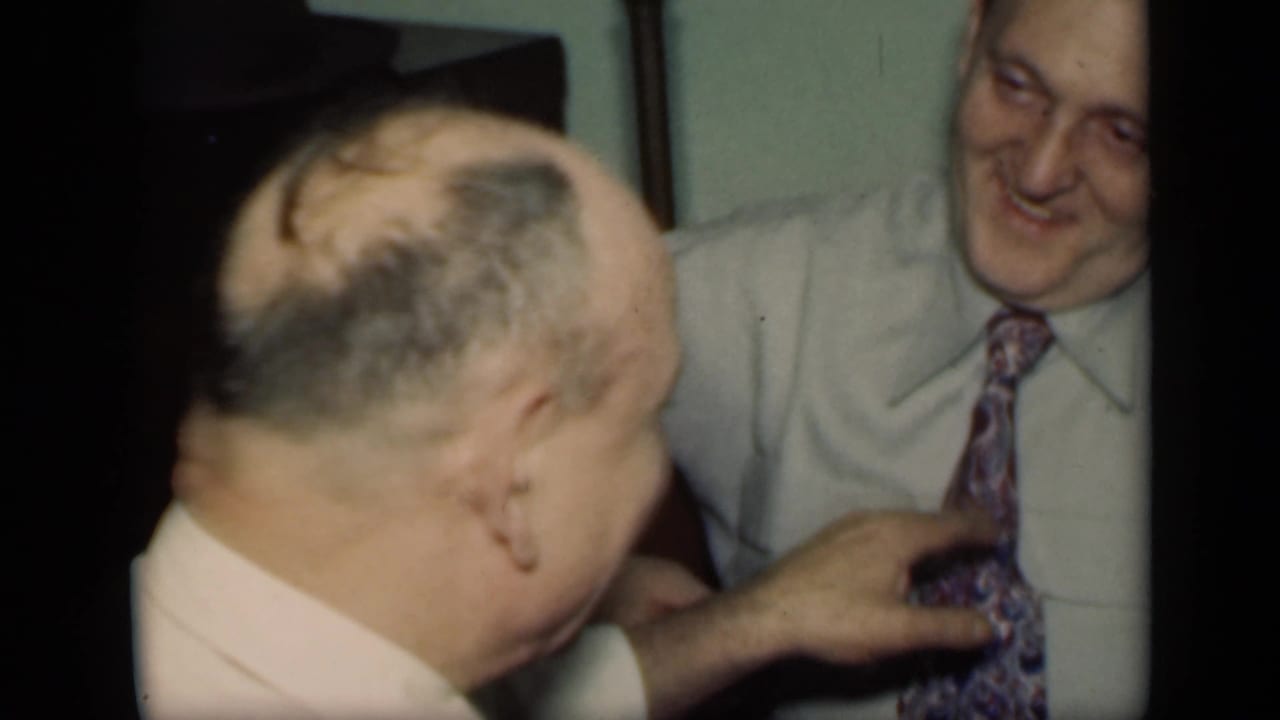 In 1949 New York home-movie footage, an older man in a pale shirt reaches toward another smiling man's patterned tie indoors.
