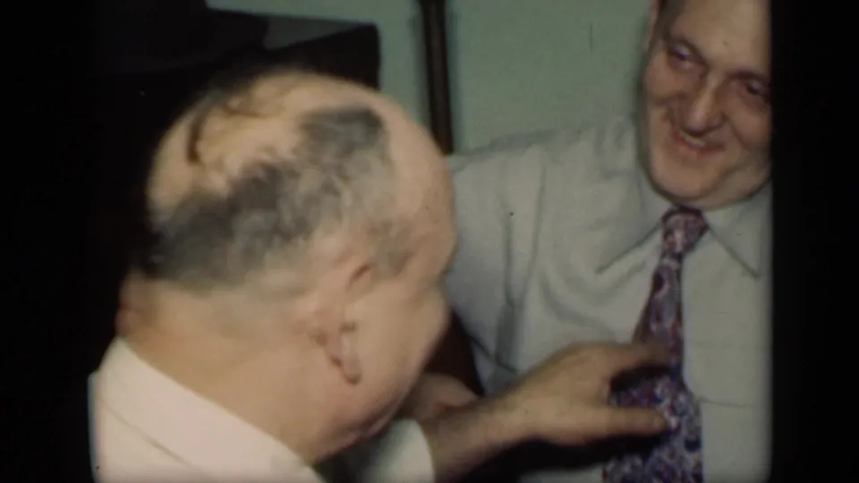 In 1949 New York home-movie footage, an older man in a pale shirt reaches toward another smiling man's patterned tie indoors.
