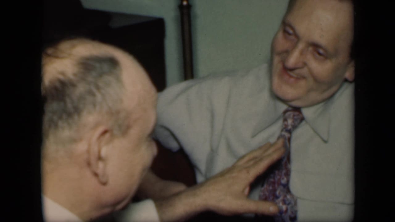 Older man smiles as another man's hand rests near his patterned tie.