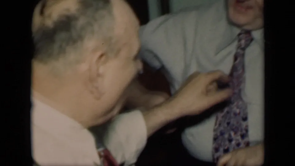 The camera stays close as one older man, seen from behind, works at another man's patterned tie in a private interior.