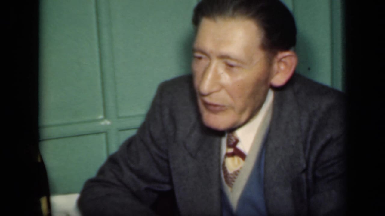 A man in a gray suit, vest, and patterned tie sits indoors against a green paneled wall in the 1949 New York home movie.