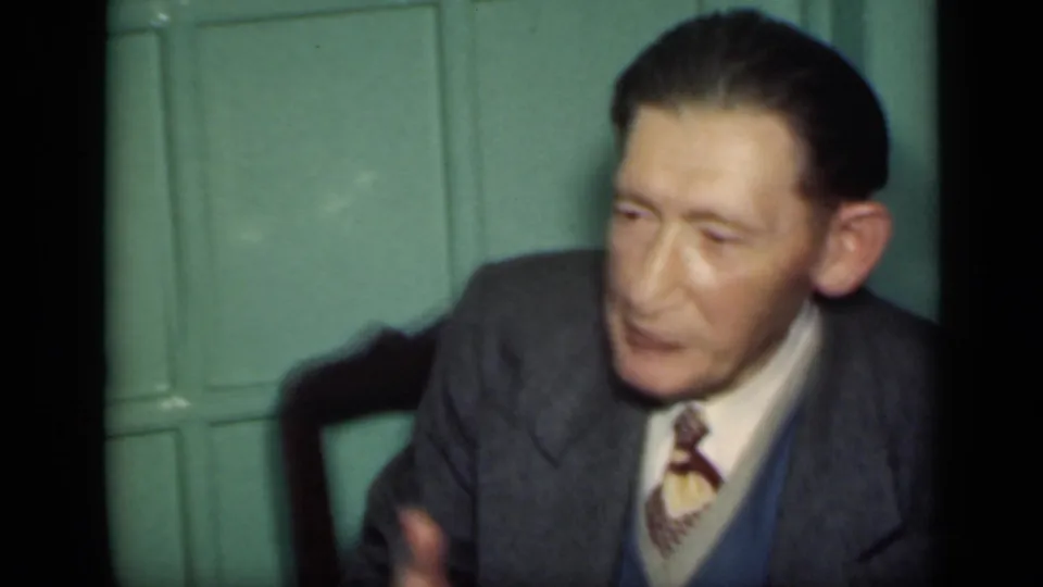 The seated man leans slightly at a table or chair edge, wearing a gray suit and tie in the 1949 New York interior scene.