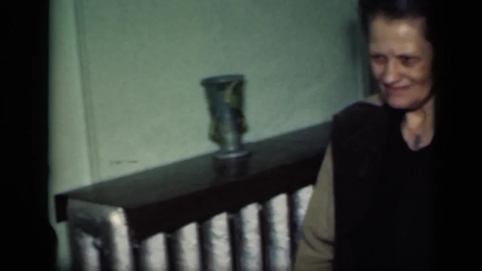 An older woman stands indoors near a dark wooden surface with a small vase, in the same 1949 New York home-movie setting.