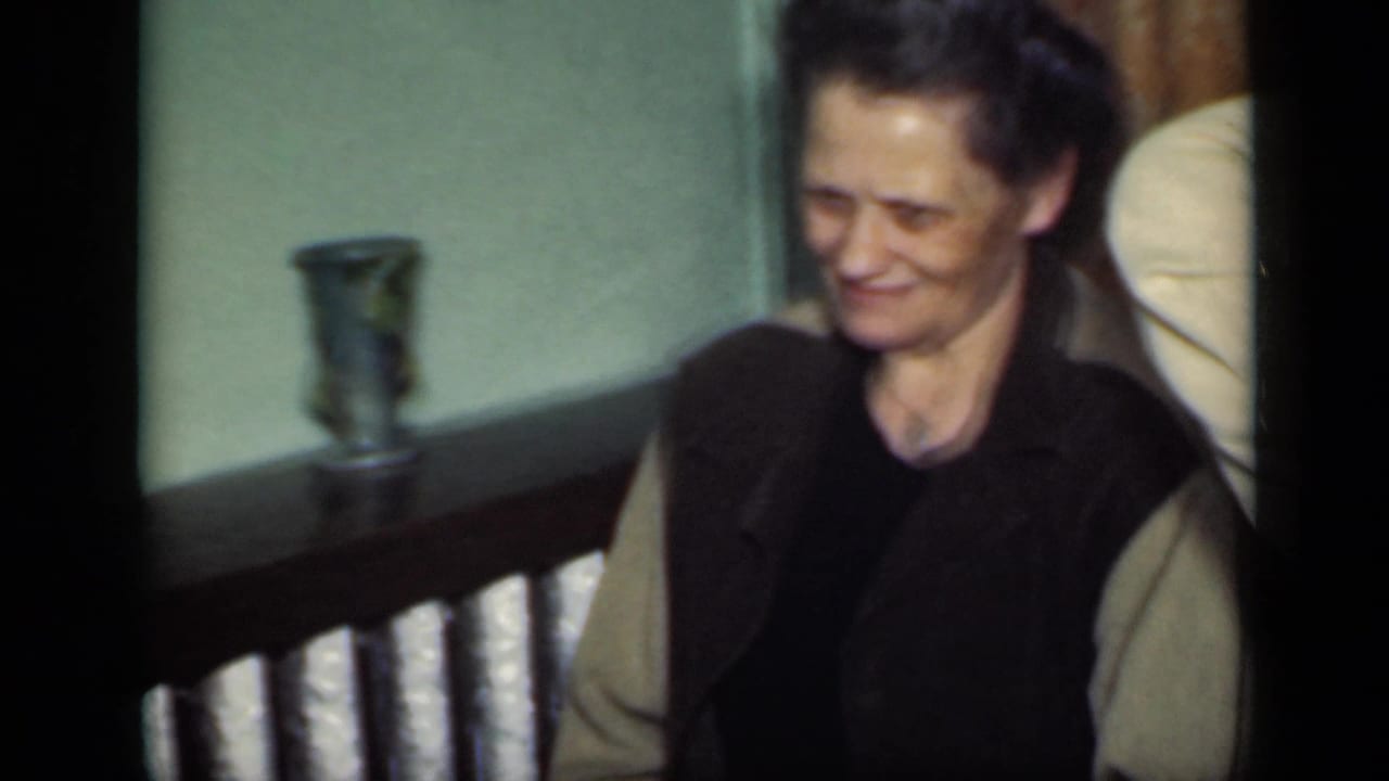 1949 New York home-movie frame showing an adult woman in a dark vest and light cardigan smiling faintly in an interior setting.